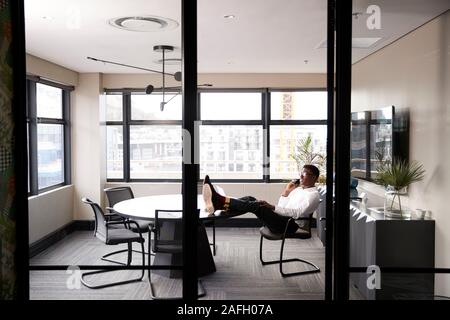 Empresario negro milenario hablando por teléfono con los pies para arriba en un escritorio, visto desde la puerta
