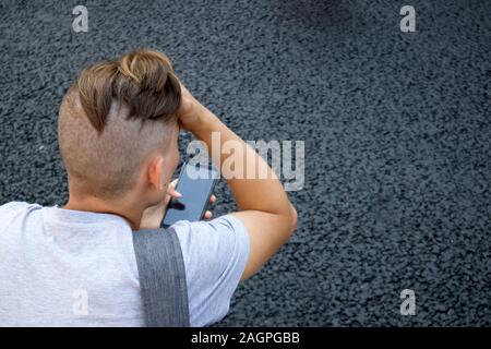 Parte posterior de la cabeza rapada con cola de caballo de joven con smartphone contra el asfalto gris Foto de stock