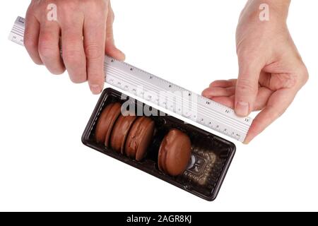 Un tecnólogo de fábrica comprueba el tamaño de donas de chocolate en una caja de plástico con una regla. Aislado en blanco studio macro Foto de stock