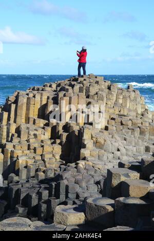 Enormes columnas de basalto de la Calzada del Gigante con una mujer en chaqueta roja de pie sobre la cumbre de tomar fotografías con su teléfono móvil, el cielo azul y