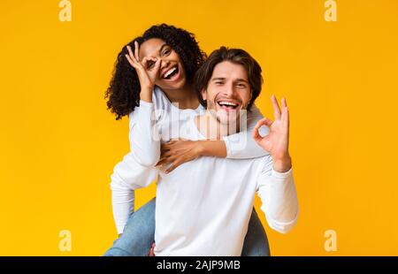 Todo está ok. Feliz pareja multirracial mostrando bien gesto mientras se divierten juntos sobre fondo amarillo, Guy aprovechar su novia afro Foto de stock