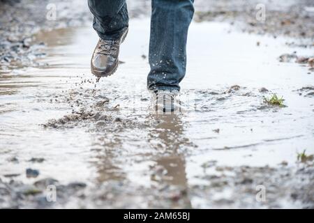 Hombre con botas de caminar marrones resistentes salpicando a través de un profundo barro embarrado en un camino después de la lluvia pesada en invierno Foto de stock