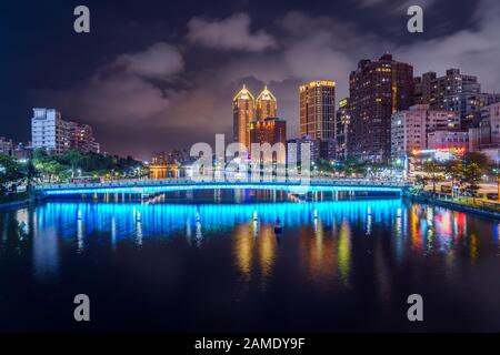 Vista increíble de un puente iluminado azul que se refleja en el agua en el río amor en Kaohsiung con el horizonte de la ciudad en el fondo por la noche Foto de stock