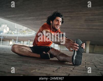 Retrato de un joven atleta sonriente en el auricular sentado bajo el puente de la ciudad estirando las piernas antes de correr