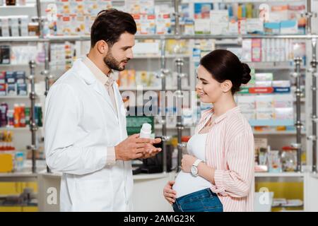 Vista lateral del farmacéutico mostrando tarro con pastillas a la mujer embarazada sonriente en boticario Foto de stock