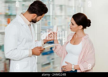 Vista lateral de la droggista con tableta digital y mujer embarazada mirando el frasco con pastillas en apotecario Foto de stock