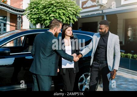 Dos hombres guapos, africanos y caucásicos, un apretón de manos, con una sonrisa mientras está de pie en la parte delantera del vehículo al aire libre. Bonita mujer caucásica está entre