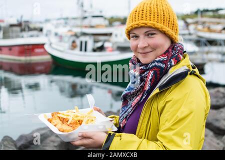 Mujer joven en amarillo impermeable come una porción de pescado y patatas fritas en el puerto de Husavik, Islandia. Fotografía de Stock.