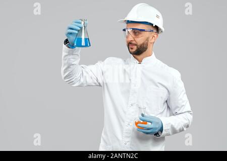 Vista frontal del químico sonriente en gafas y casco que sostiene dos matraces con líquido azul y naranja. Científico en el laboratorio blanco bata haciendo experimento en laboratorio, aislado en gris. Concepto de química. Foto de stock
