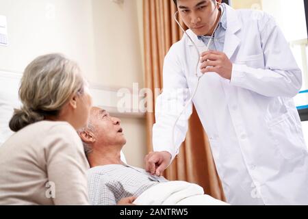 Paciente varón anciano asiático acostado en la cama que está siendo examinado por el joven doctor en el hospital