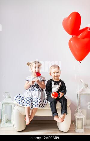 Niña y Niño sentado en una silla blanca cerca de globos con forma de corazón. Chica lamiendo un círculo rojo. Día de San Valentín concepto
