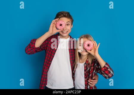 Felices hermanos que cubren los ojos con donuts, posando juntos sobre fondo azul Foto de stock