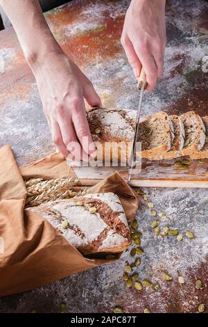 Las manos femeninas de inyección vertical cortan pan de masa cocida recién horneado con semillas de girasol y calabaza en una servilleta marrón. Orejas de trigo.