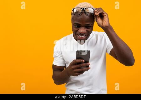 Guapo africano de piel oscura con gafas con poca vista sonriente mirando el teléfono sobre un fondo amarillo. Foto de stock
