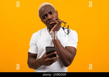 Guapo africano de piel oscura con pelo blanco en gafas con vista pobre cuidadosamente sostiene un teléfono en sus manos sobre un fondo de estudio amarillo con Foto de stock