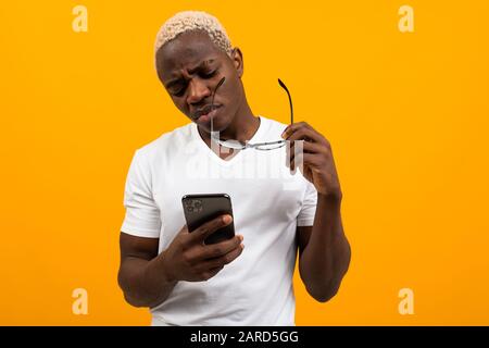 Guapo africano de piel oscura con pelo blanco en gafas con vista pobre mira cuidadosamente el teléfono en un fondo amarillo. Foto de stock