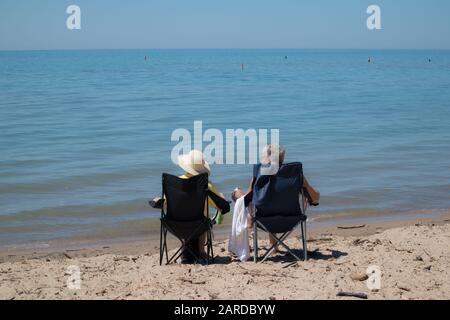 Una pareja madura, probablemente retirada, mirando el lago Huron mientras se sienta en su silla plegable en la playa. Foto de stock