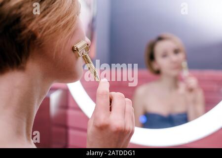 La joven mujer massando su cara con gua sha spiked rodillo de metal en su cuarto de baño Foto de stock
