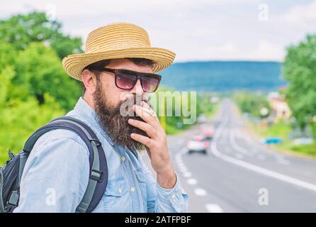 Viejo hábito. Hombre con barba y bigote en sombrero de paja fumar cigarrillo, carretera fondo defocused. Viajero elegante hipster tomar freno con cigarrillo. Fumar cigarrillos antes de un largo viaje.