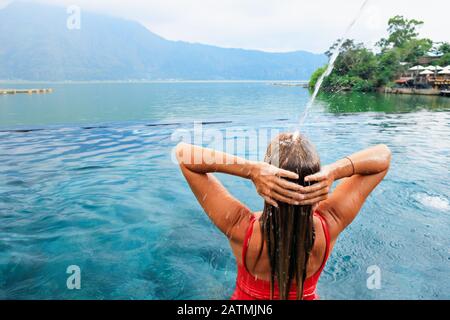 Una mujer joven se relaja bajo el agua corriente en la piscina infinita con hermosas vistas al lago. Balneario natural de aguas termales en el volcán Batur. Visita turística a Kintamani