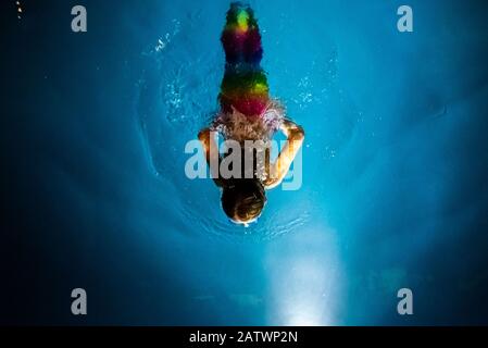 Niña disfrazada de sirena buceando hacia la luz de una piscina azul, con un fondo de ensueño de aguas cristalinas.