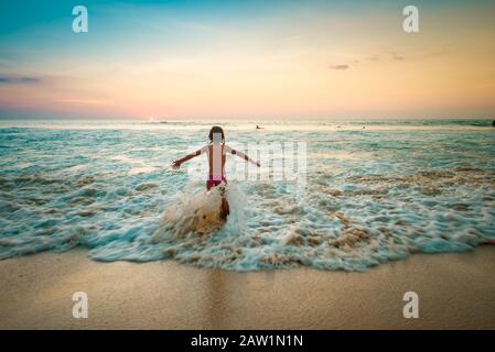 Niña jugando en una playa tropical en el borde de aguas claras con puesta de sol y cielo azul jugando en el agua con las olas viniendo.