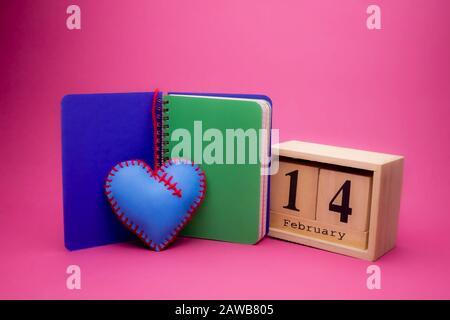 Día de San Valentín todavía vida con mano cosida corazón azul textil, cuaderno y fecha de madera sobre un fondo rosa Foto de stock