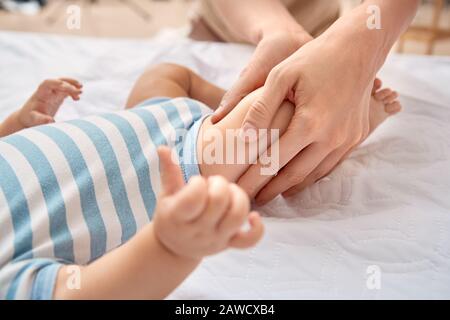 Cuidado Del Bebé. Madre joven haciendo masaje pediátrico masajeando las piernas de un hijo pequeño acostado en la cama de cerca
