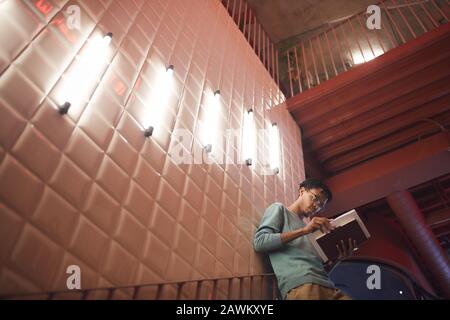 Retrato en gran angular de un joven afroamericano que sostiene libros mientras se inclina en la pared en el campus universitario, espacio para copias
