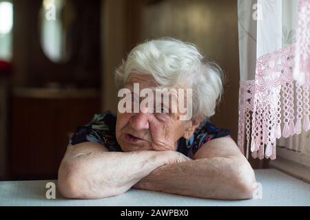 Una vieja mujer se sienta pensadamente en la mesa. Foto de stock
