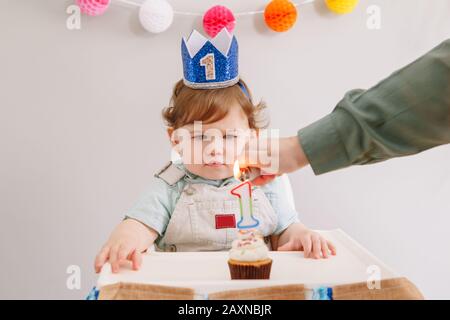 Foto Lindo bebé caucásico sonriente en corona azul celebrando su primer  cumpleaños en casa. Niño niño pequeño sentado en silla alta comiendo  sabroso postre de magdalenas con la palabra topper Uno. Concepto