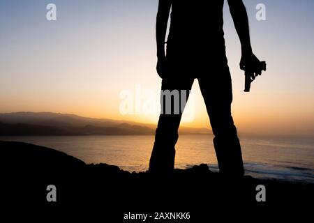 Silueta del hombre sosteniendo una pistola al atardecer en los acantilados con vistas al océano