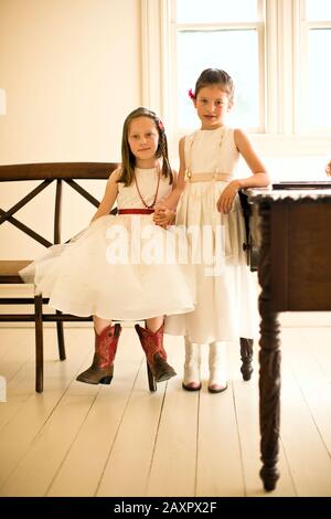 Las niñas jóvenes en vestidos blancos de fiesta y botas de vaquero tienen  las manos mientras una se sienta en un banco de madera y su amiga se  encuentra junto a ella