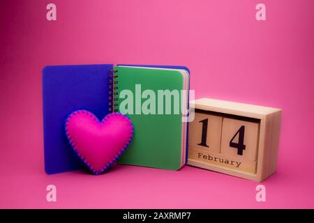Día de San Valentín todavía vida con mano cosida corazón rosa textil, cuaderno y la fecha de madera sobre un fondo rosa Foto de stock