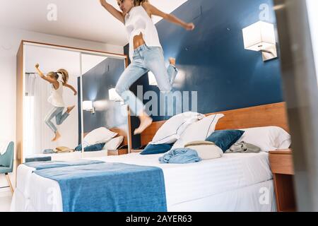 Chica rubia saltando sobre una cama de hotel con las sábanas azules y un espejo Foto de stock