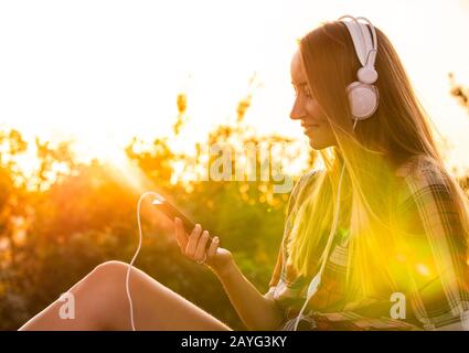 Feliz joven rubia mujer con pelo largo y ojos azules escuchando música con headpones blancos sentados afuera al atardecer.