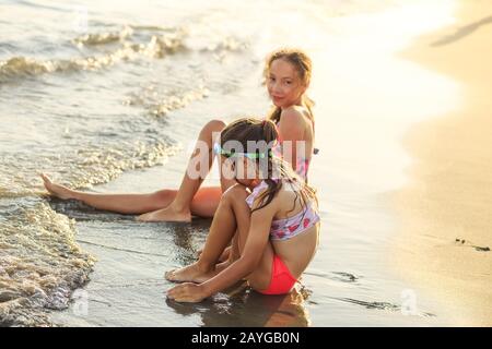 Dos pequeñas chicas del cute sentadas en el mar y jugando con las olas al atardecer. Verano Sunny Day, Costa Del Océano, Happy Kids Concept