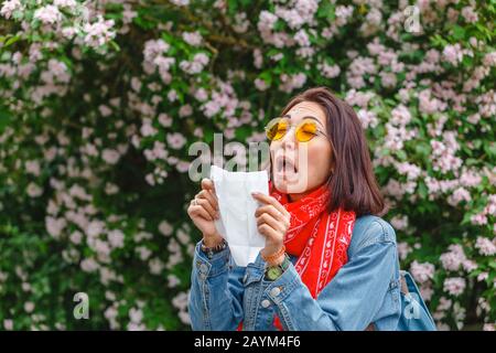 Concepto de alergia. Una joven mujer asiática estornuda y sopla su nariz con un pañuelo y sufre en la primavera entre árboles florecientes y florecientes. Foto de stock