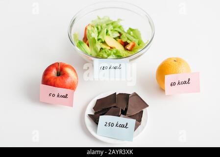 Frutas frescas, chocolate y ensalada con calorías en tarjetas de fondo blanco, dieta de recuento de calorías Foto de stock