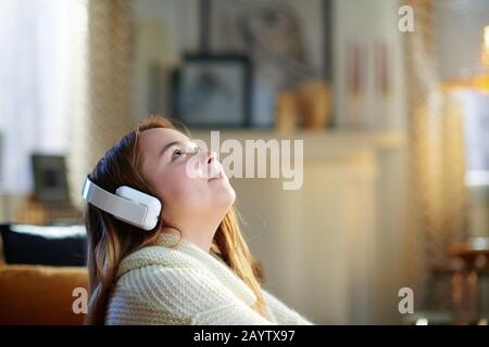 mujer joven moderna con pelo rojo en un suéter blanco escuchando música con auriculares y mirando hacia arriba en el espacio de copia mientras se sienta cerca del sofá en el modo Foto de stock