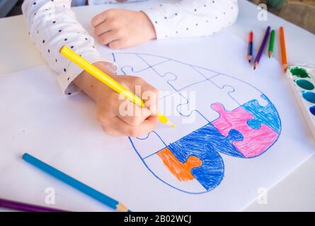 Un niño está dibujando un corazón colorido con rompecabezas sobre fondo blanco como símbolo de autismo, con lápices de colores y acuarelas. Foto de stock