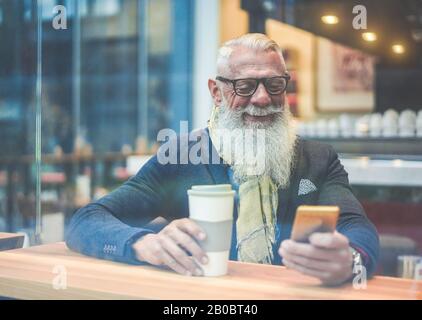 Hombre de negocios sénior que utiliza la aplicación para smartphone mientras bebe café en el bar del salón - empresario hipster que desayuna con capuchino - Trabajo, moda