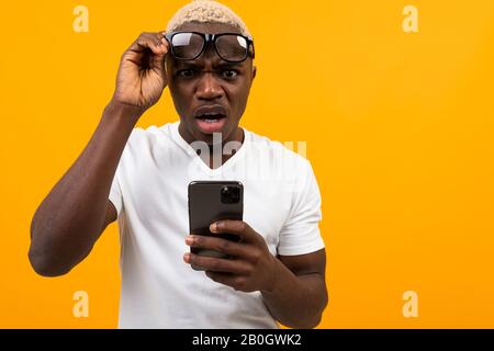 Guapo africano de piel oscura en gafas con mala vista parece sorprendido en el teléfono sobre un fondo amarillo con espacio de copia Foto de stock