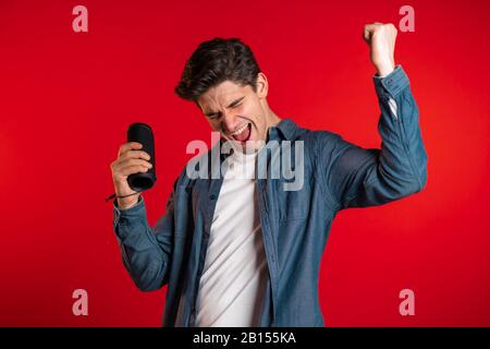 Hombre sonriendo, bailando con altavoz portátil inalámbrico en el estudio sobre fondo rojo. Música, concepto de danza. Foto de stock