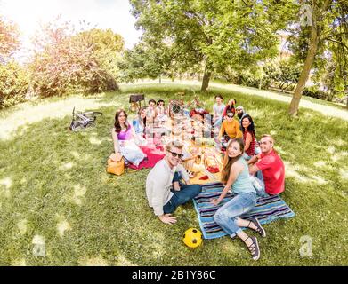 Grupo de amigos felices haciendo picnic en el parque público al aire libre - jóvenes bebiendo vino y riendo en la naturaleza - enfoque principal en los chicos de fondo - Juventud a Foto de stock