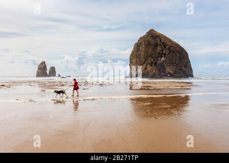 Mujer joven caminando su perro en la playa en Cannon Beach, Oregon, Estados Unidos con la famosa Haystack Rock en el fondo.
