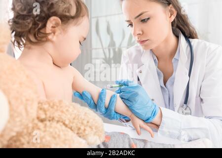Una joven pediatra realiza una vacunación de una niña pequeña. La chica sostiene una mascota. Foto de stock
