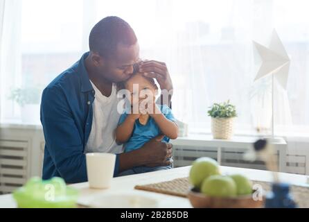 Retrato de un feliz padre africano besando a un pequeño hijo en la frente mientras se sienta en la cocina iluminada por el sol, espacio de copiado