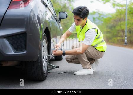 Hombre asiático joven con chaleco verde de seguridad cambiando el neumático perforado en su coche aflojando las tuercas con una llave de rueda antes de levantar el vehículo con el gato