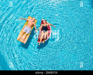 Por encima de la vista vertical de la gente pareja mayor tomando las manos con amor y divertirse en la piscina azul claro juntos disfrutando de las vacaciones de verano con el moderno color lilos colchón inflable